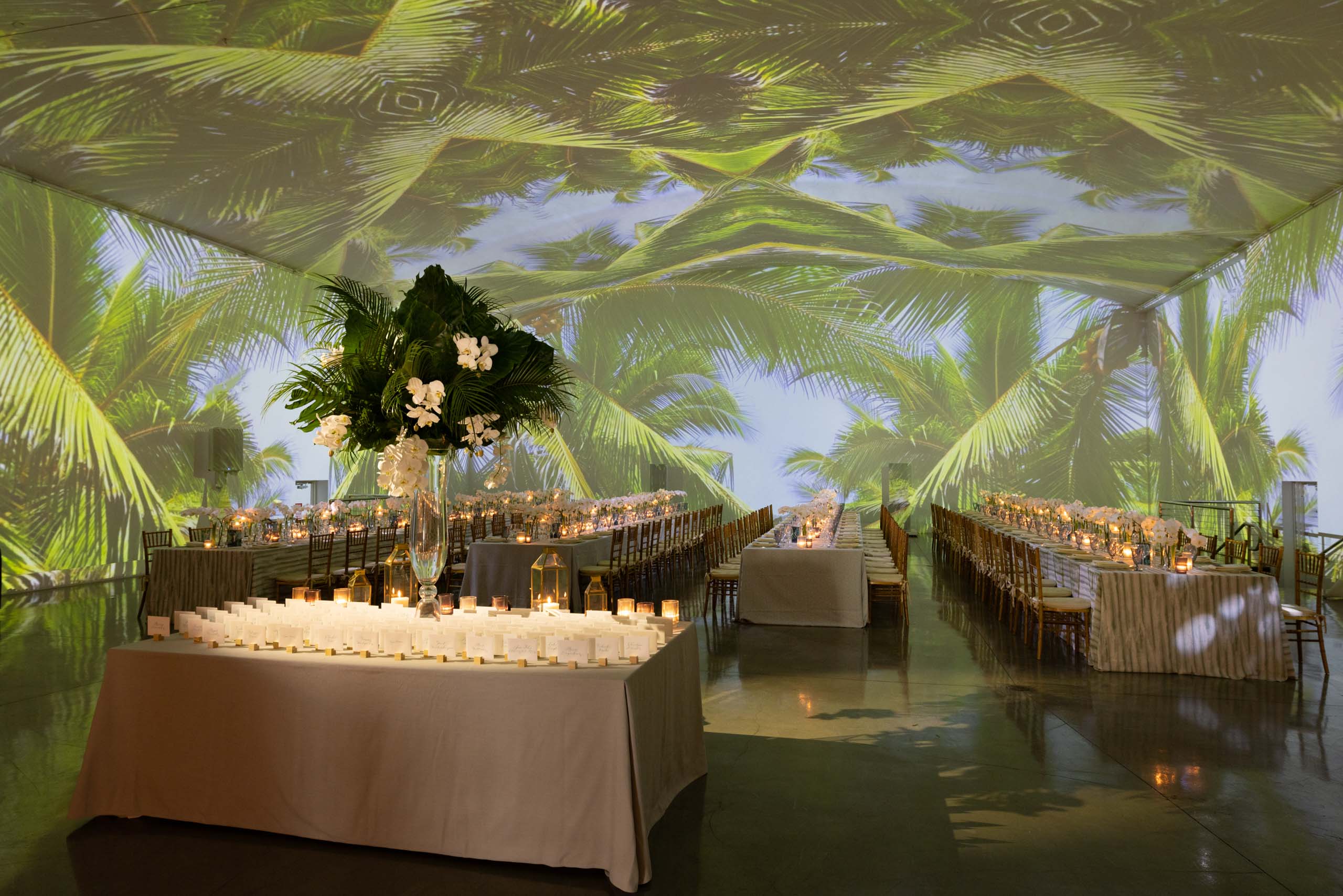 Elegant event venue space with tropical palm projections on the ceiling, candlelit tables, and a floral centerpiece.