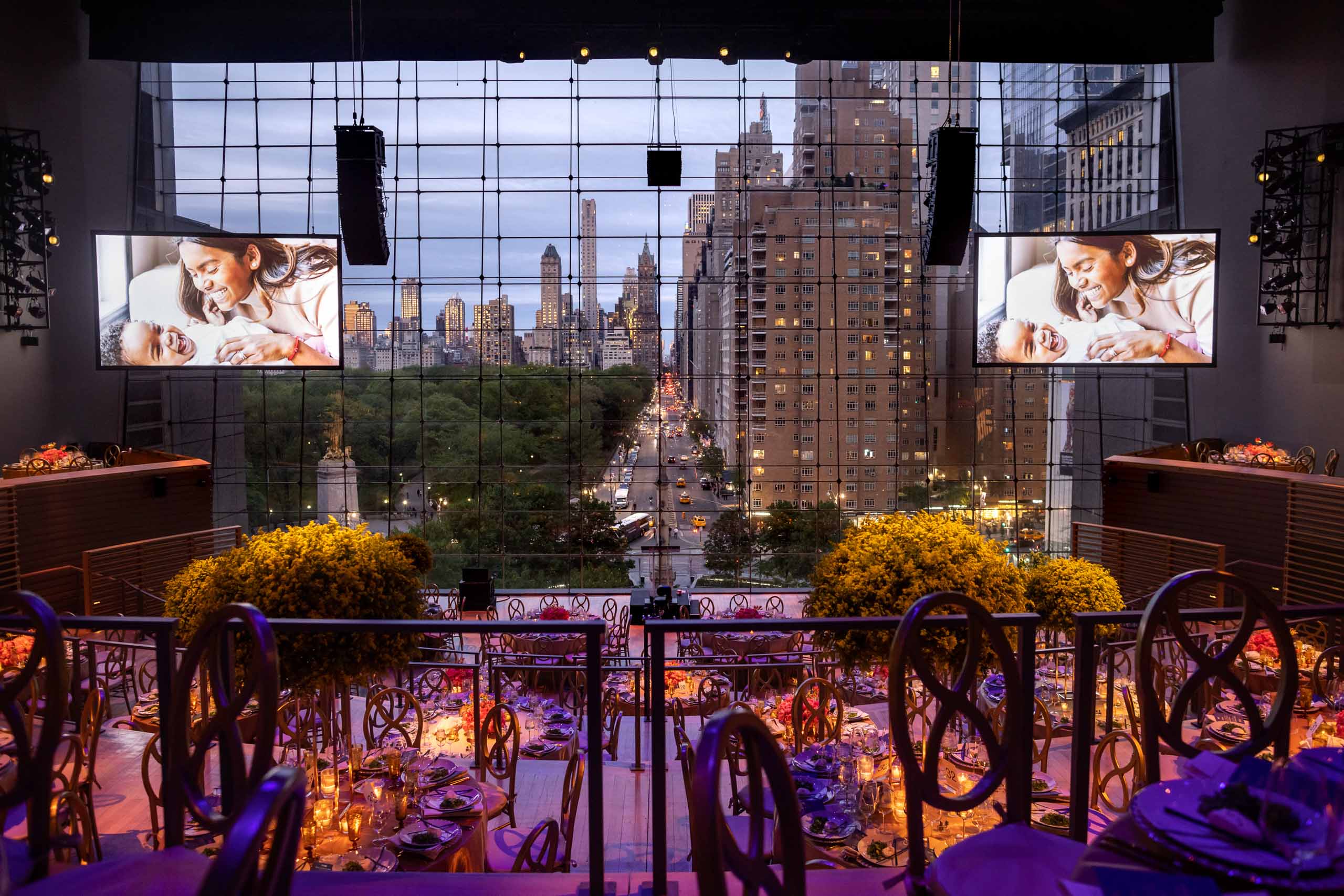 An elegant event venue space with large video screens showing images of a mother and child, overlooking an urban skyline at dusk, adorned with floral arrangements and set tables.